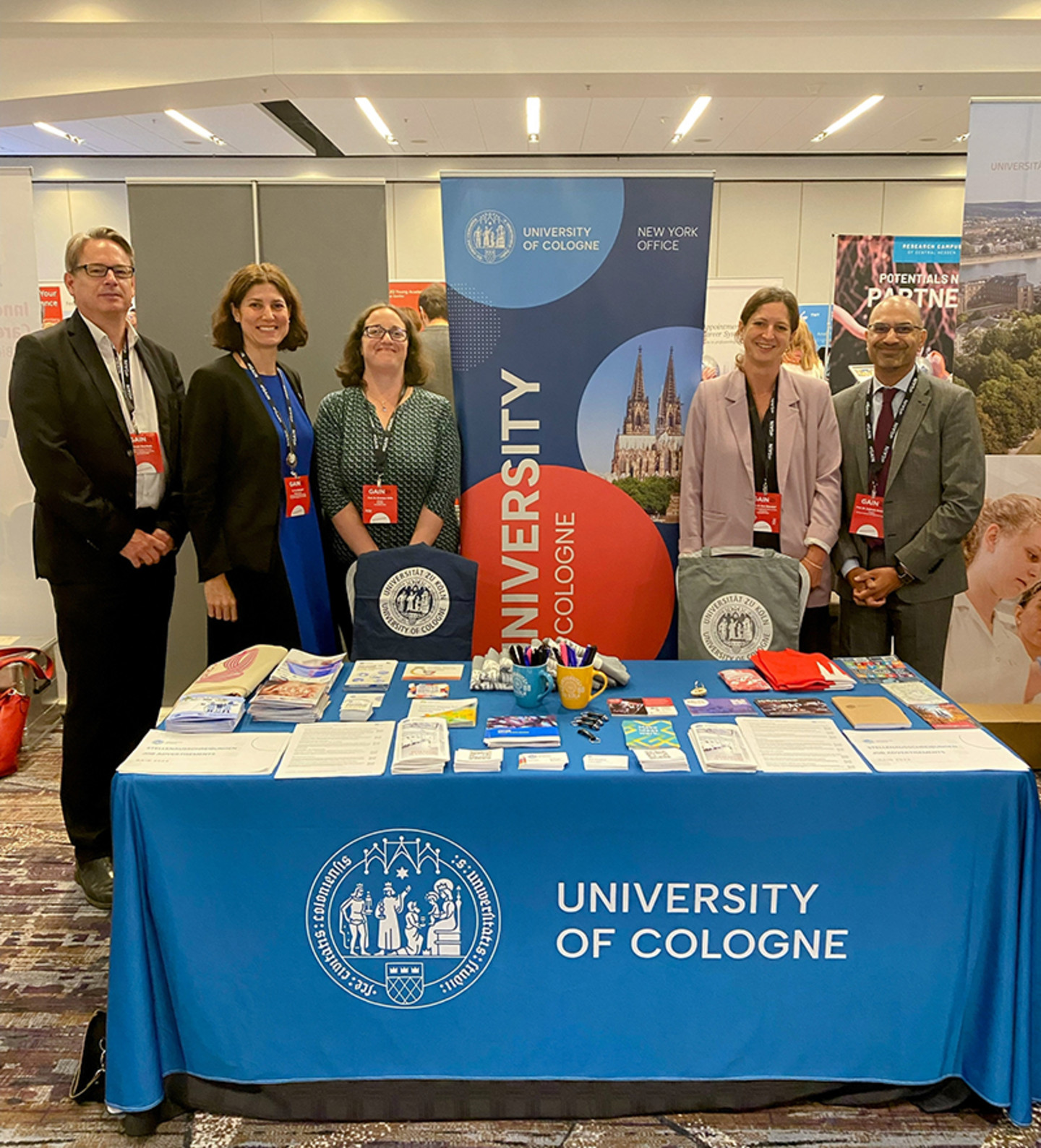 Two men and three women standing in front of a table with various information materials, smiling and looking at the camera. The logo of the University of Cologne can be seen in the background and on the tablecloth.