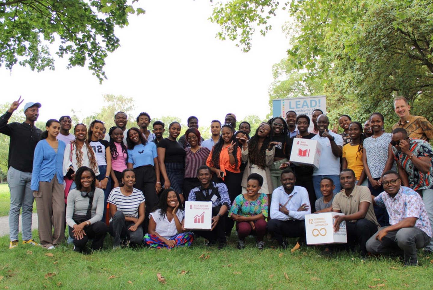 Gruppenfoto mit den LEAD!-Stipendiaten und Dr. Georg Verweyen (rechts) vom International Office auf einer Wiese im Universitätspark. Einige halten Pappwürfel mit den UN-Zielen für nachhaltige Entwicklung auf jeder Seite. Zu sehen sind SDG 4: Hochwertige Bildung, SDG 8: Menschenwürdige Arbeit und Wirtschaftswachstum und SDG 12: Nachhaltiger Konsum und Produktion. Im Hintergrund ist ein „LEAD!“-Banner zu sehen.  Übersetzt mit DeepL.com (kostenlose Version)